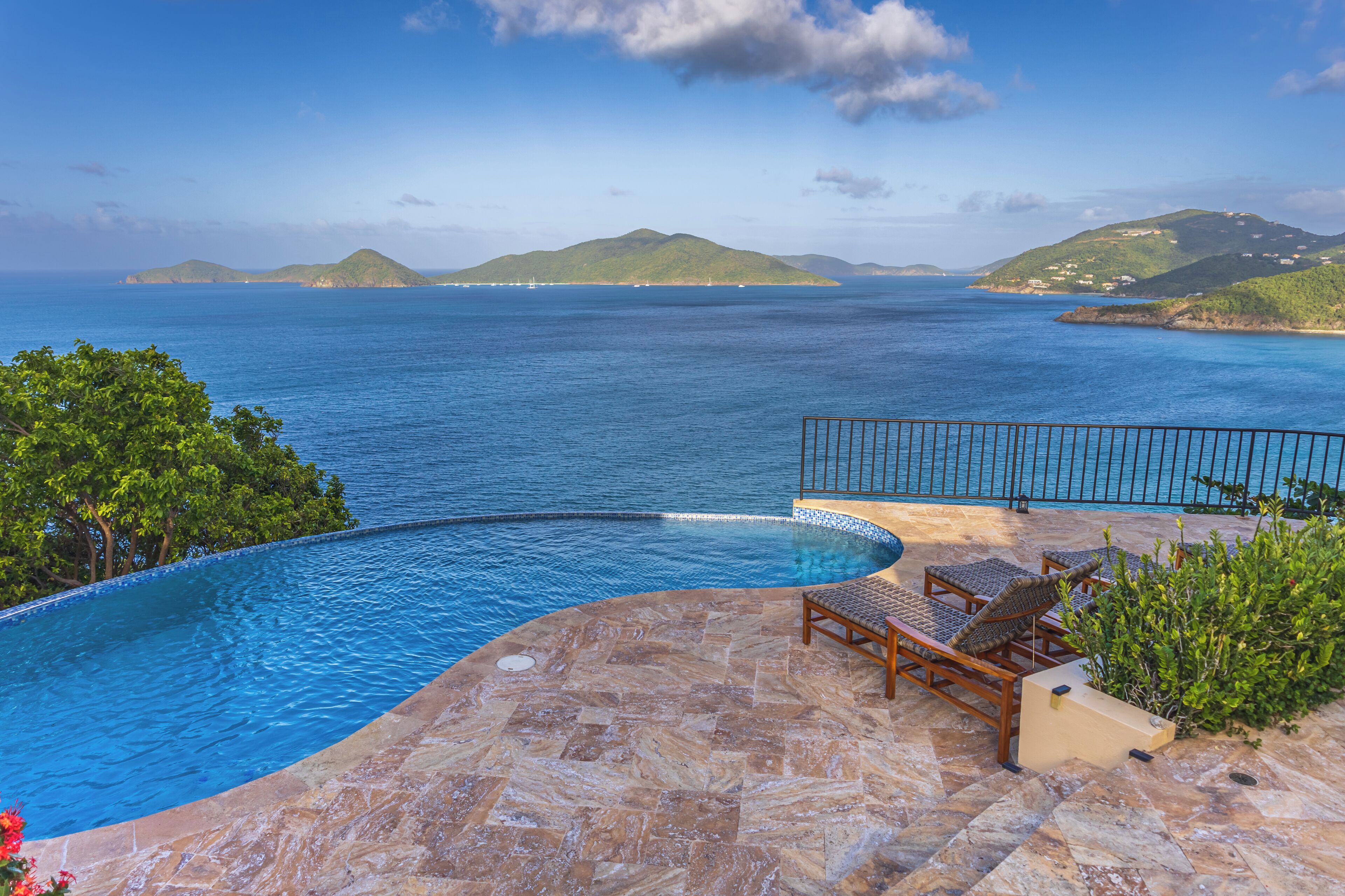 A infinity pool with a view of the sea and hills, outdoor seating, and a stone patio.