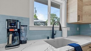 A kitchen with a coffee maker, a sink, and a window with a view of a car and trees. - Blanchet Casita – Stylish 1BR Casita w/ Fire Pit & Patio (Vancouver)