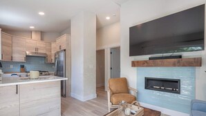 A modern kitchen with light wood cabinets, a stainless steel refrigerator, and a built-in oven. There is a seating area with a brown leather chair and a coffee table. A flat-screen TV is mounted above a fireplace with a blue-tiled surround. - Blanchet Casita – Stylish 1BR Casita w/ Fire Pit & Patio (Vancouver)