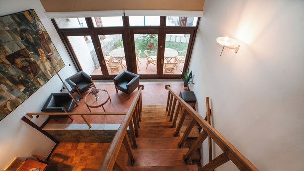 A spacious living area with a wooden staircase, a dining table with chairs, and a large window overlooking a garden. - Casa d'Abreu Neto - Siza's First Work (Matosinhos)