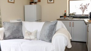 A modern living room with a white sofa, grey cushions, a white blanket, a white refrigerator, a kitchen area with a sink and window, and framed pictures on the wall. - Lannlyvri Lodge (Bodmin)