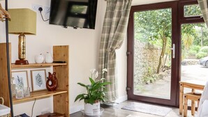 A cozy room with a television mounted on the wall, a wooden shelf with decorative items, a potted plant, and a view of a garden through a glass door. - Lannlyvri Lodge (Bodmin)