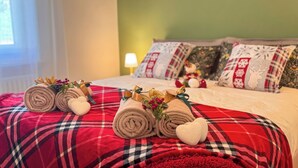 Un letto rifatto con cura, asciugamani arrotolati e cuscini a forma di cuore, una coperta a quadri e cuscini decorati con motivi a fiocchi di neve e renne.