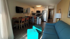Ein modernes Wohnzimmer mit einem blauen Sofa, einem Essbereich mit Holzsesseln und einer Küche mit weißen Schränken und einer Mikrowelle.
