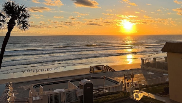 Blick auf einen Strand mit Sonnenuntergang, einer Palme und einem Parkplatz mit einem Auto.