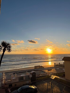 A beach view with a sunset, a palm tree, and a parking area with a car. - Bike Week 2026...Daytona Oceanfront 1BR Condo - Full Kitchen - Sleeps 4 (Daytona Beach Shores)