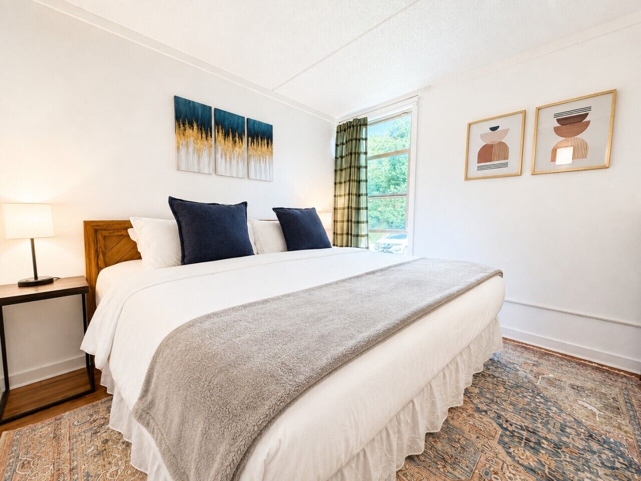 Une chambre à coucher comprenant un lit, une table de chevet, une fenêtre avec des rideaux et deux œuvres d’art accrochées au mur.
