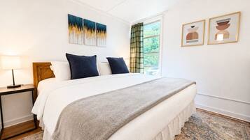 A bedroom with a bed, a nightstand, a window with curtains, and two wall-mounted artworks.