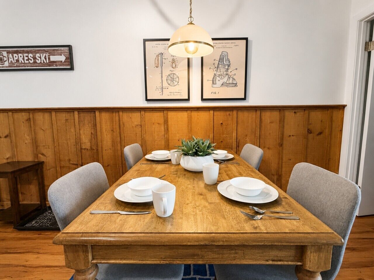 Un coin repas avec une table en bois dressée pour quatre personnes, un luminaire suspendu et des reproductions encadrées de dessins de brevets accrochées au mur.