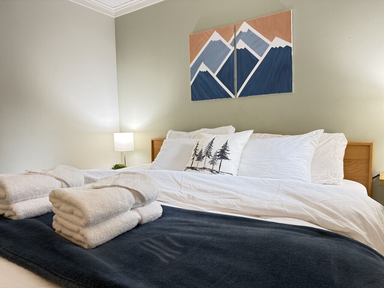Ein ordentlich gemachtes Bett mit weißen Leinentüchern, einer dunklen Tagesdecke und einem Kopfkissen mit Waldmuster. Auf dem Bett liegen gefaltete Handtücher, darüber hängt ein Wandbild.