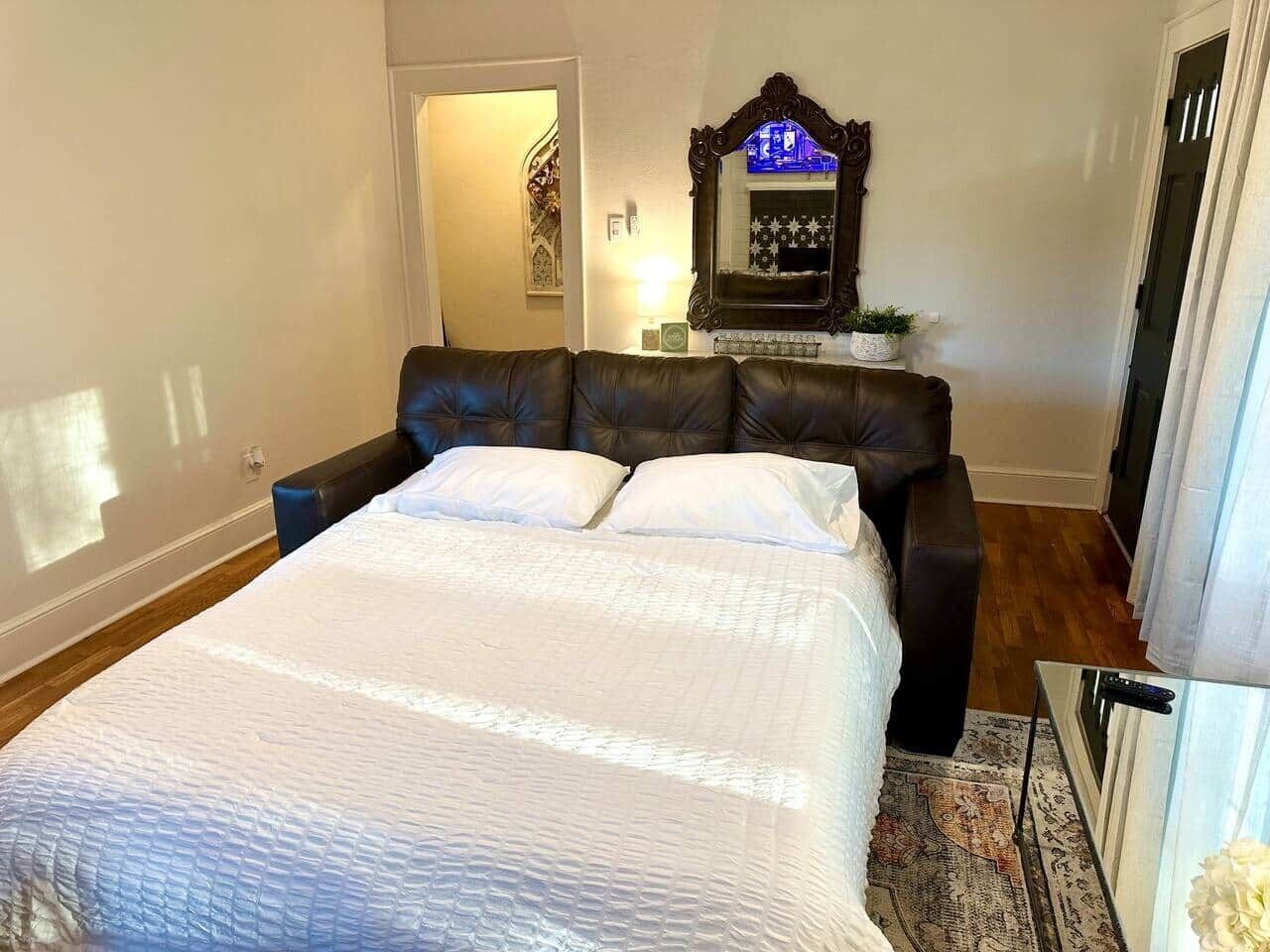 A bedroom with a large bed, a dark leather sofa, a small table with a lamp, and a mirror on the wall.