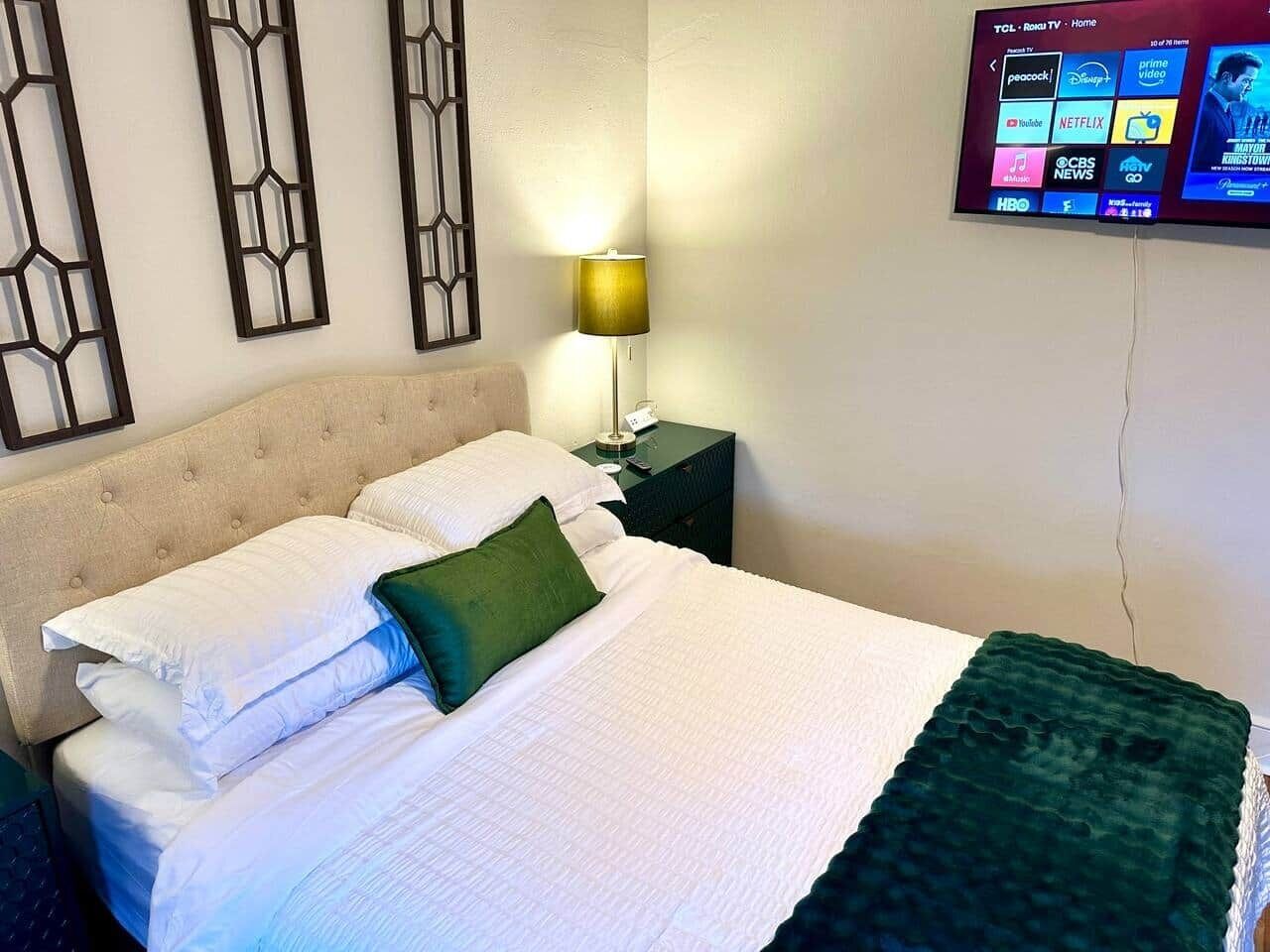 A neatly made bed with white bedding and a green pillow, a bedside lamp, and a TV displaying various channels.