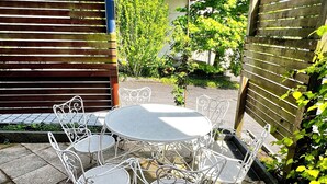 Eine Terrasse mit rundem Tisch und weißen Metallstühlen, umgeben von Steinplatten.