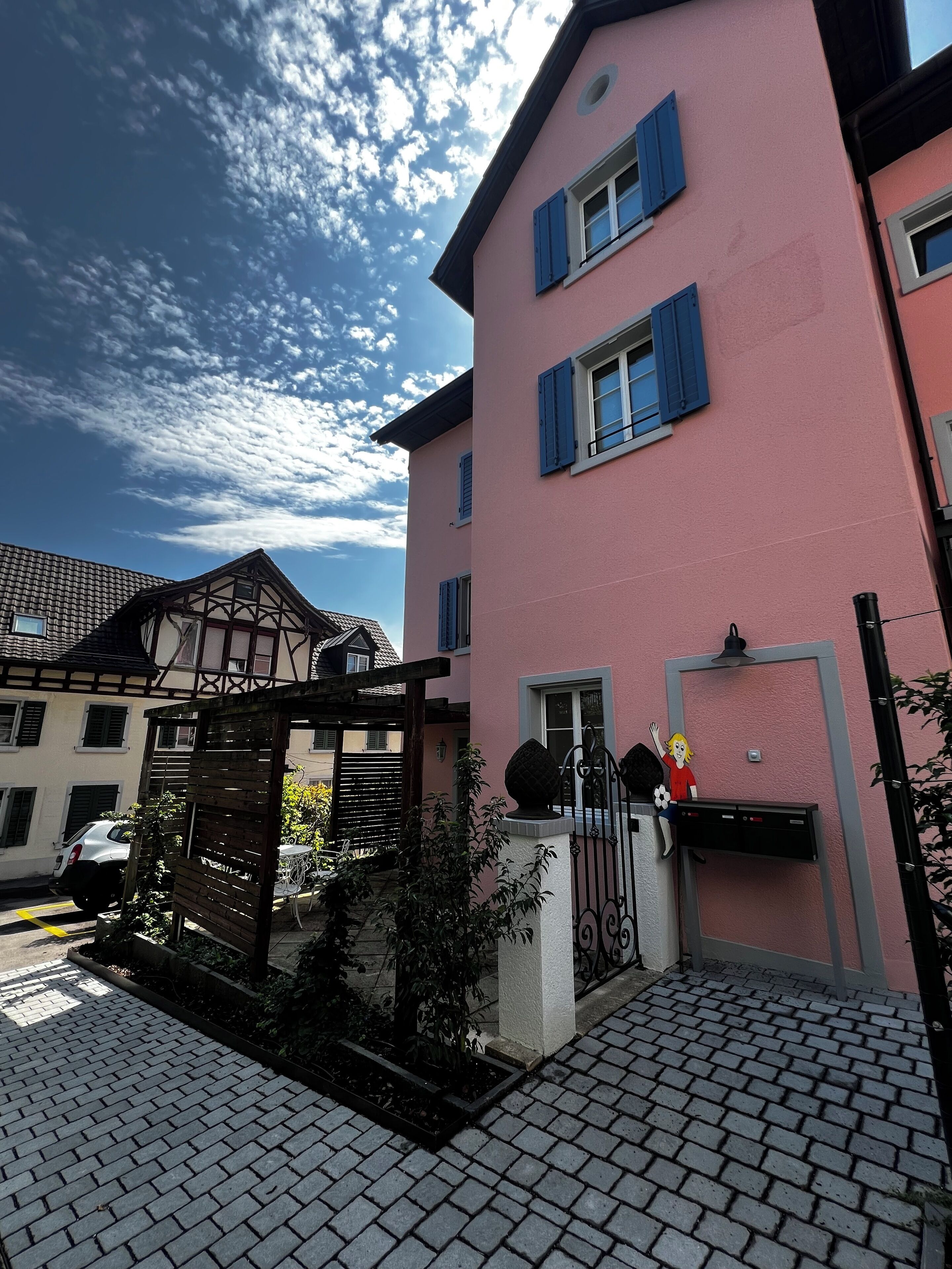 A pink building with blue shutters, a wooden pergola, and a paved entrance.