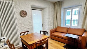 Ein Wohnzimmer mit einem Holzespeisetisch, Stühlen, einem braunen Ledersofa, einem kleinen schwarzen Beistelltisch und einem Fenster mit Vorhängen.