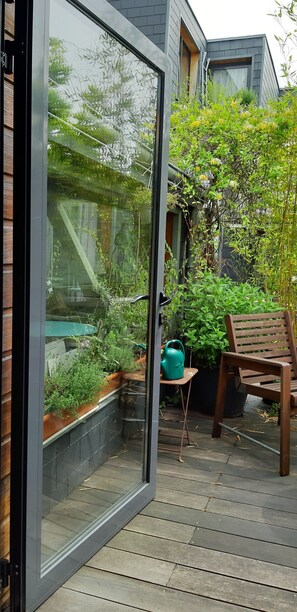 Uma porta de vidro dá acesso a um pátio com um banco de madeira, um vaso de planta e uma mesa pequena.