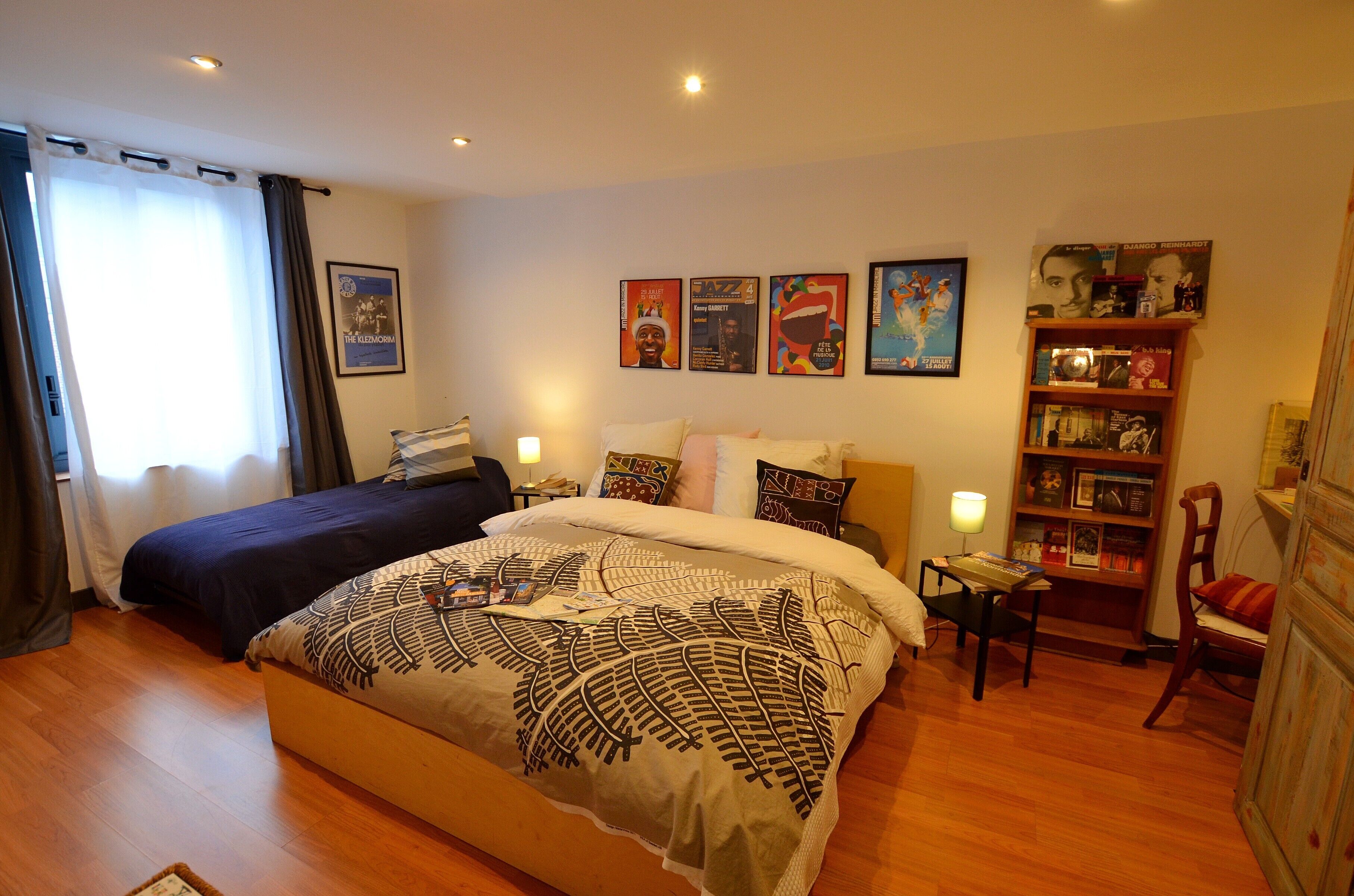 Een tweepersoonsbed met een bedsprei met dessin, een nachtkastje met lamp, een boekenkast en een raam met gordijnen.