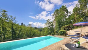En swimmingpool med krystalklart, blåt vand, omgivet af stentøj og frodig vegetation. Liggestole og et bord med en flaske og et glas.