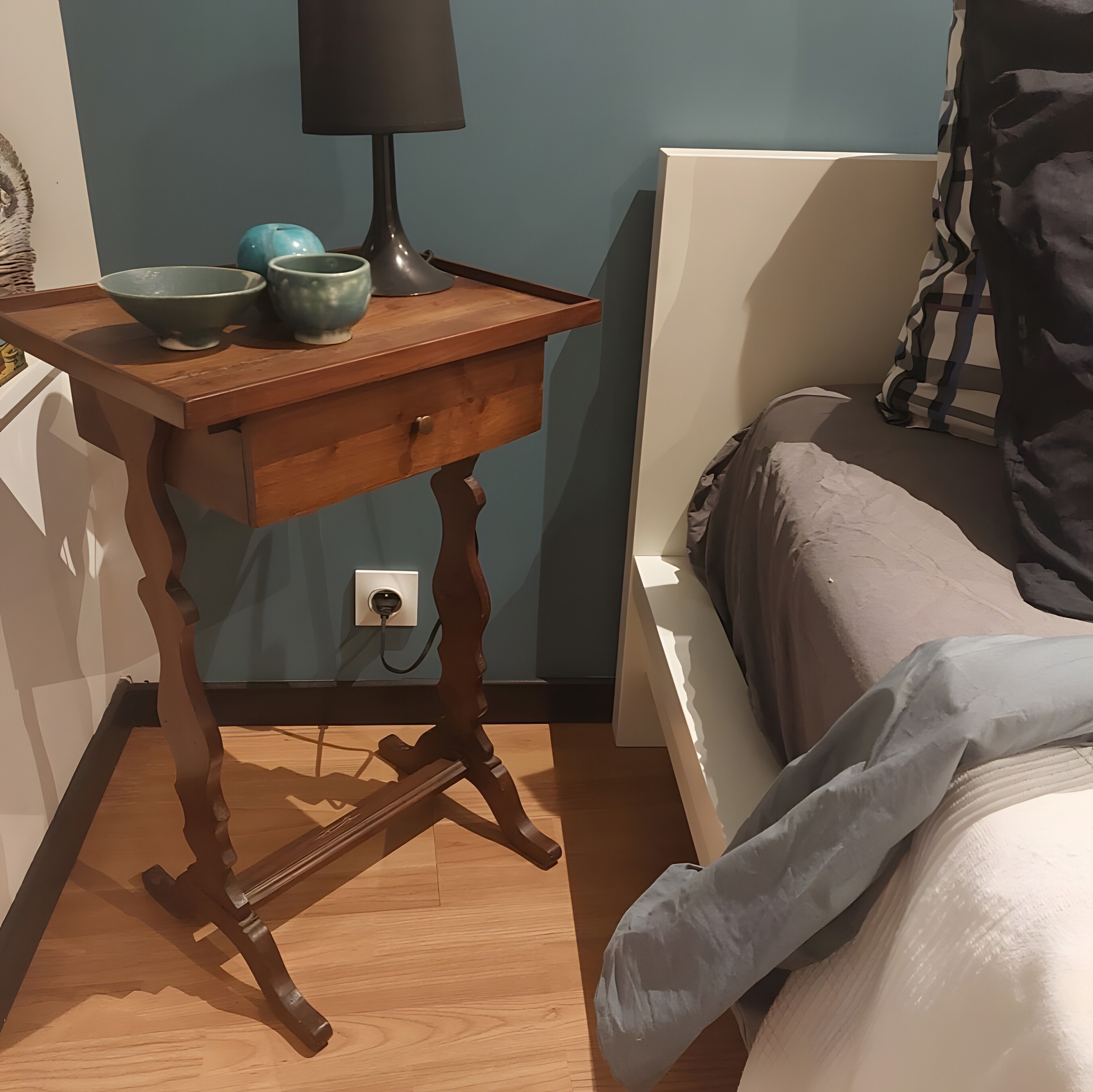 Une chambre à coucher avec une table de chevet en bois, une lampe, un lit et une prise électrique au mur.