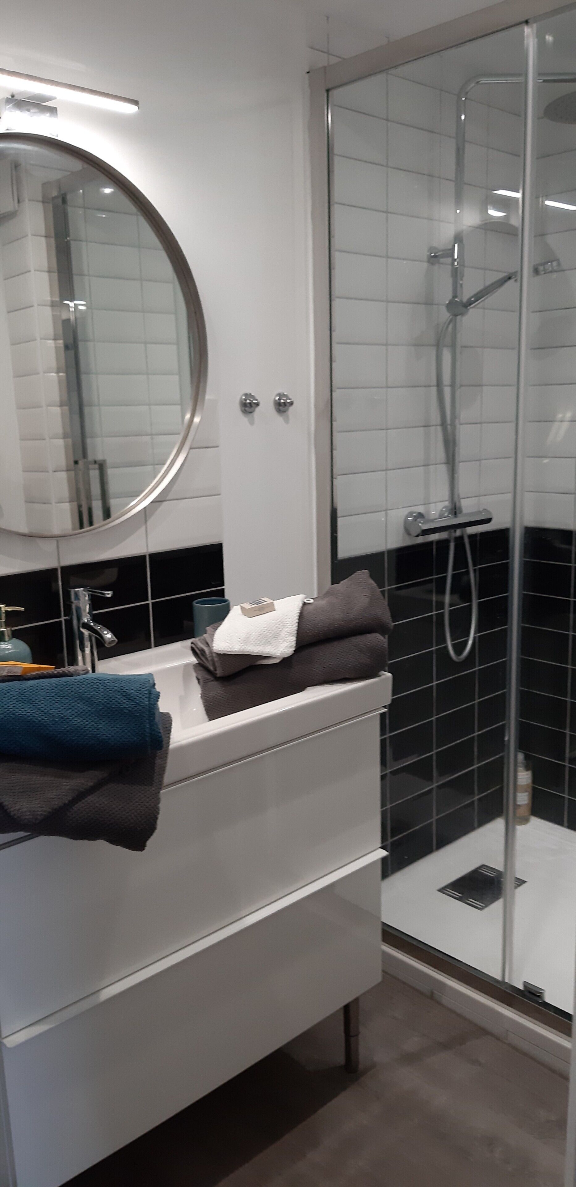 A modern bathroom with a round mirror, a white vanity, a glass shower enclosure, and a black tiled wall.