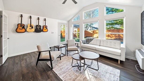 Ett rymligt vardagsrum med en vit soffa, två fåtöljer, ett soffbord och en takfläkt. På väggen hänger fyra gitarrer, och stora fönster släpper in naturligt ljus och ger utsikt över omgivningen.