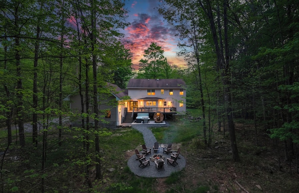 A house surrounded by forest with a fire pit and outdoor seating.