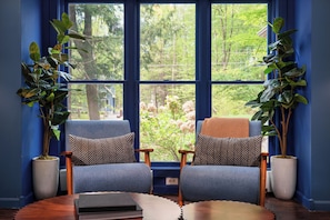 A room with two armchairs, a round table, and large windows with a view of trees and a house.