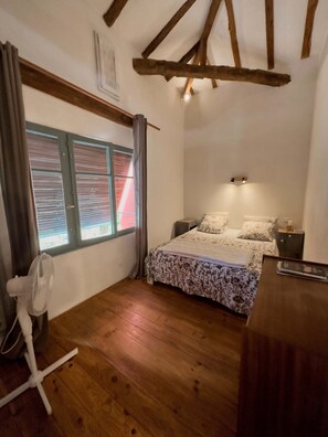 Une chambre avec un plancher en bois, un lit avec une literie à motifs floraux, un bureau, une chaise et une fenêtre avec des stores.