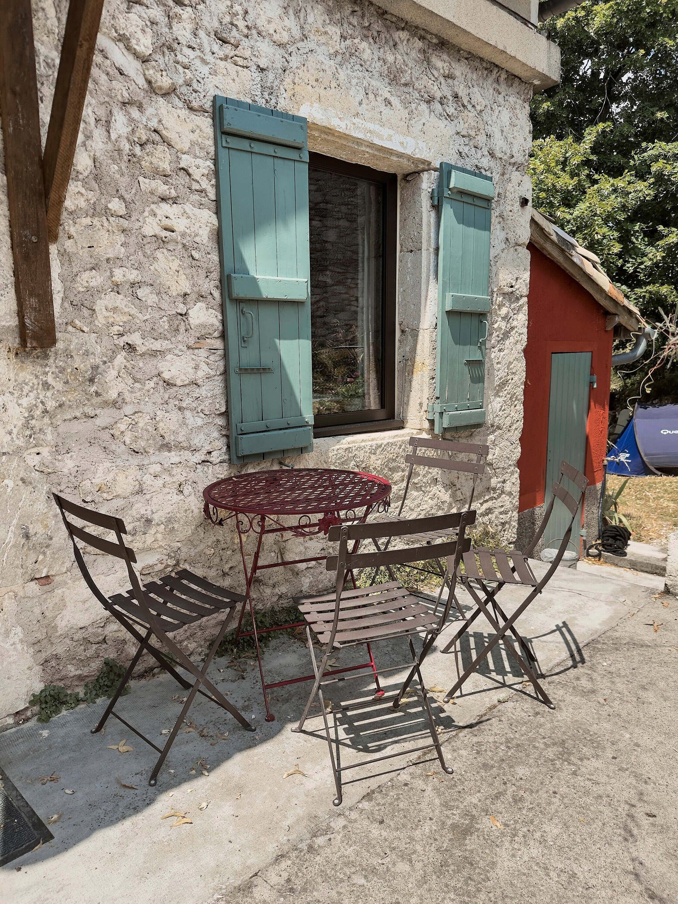 A rustic outdoor seating area with a metal table and chairs, a stone wall, and a small window with shutters.