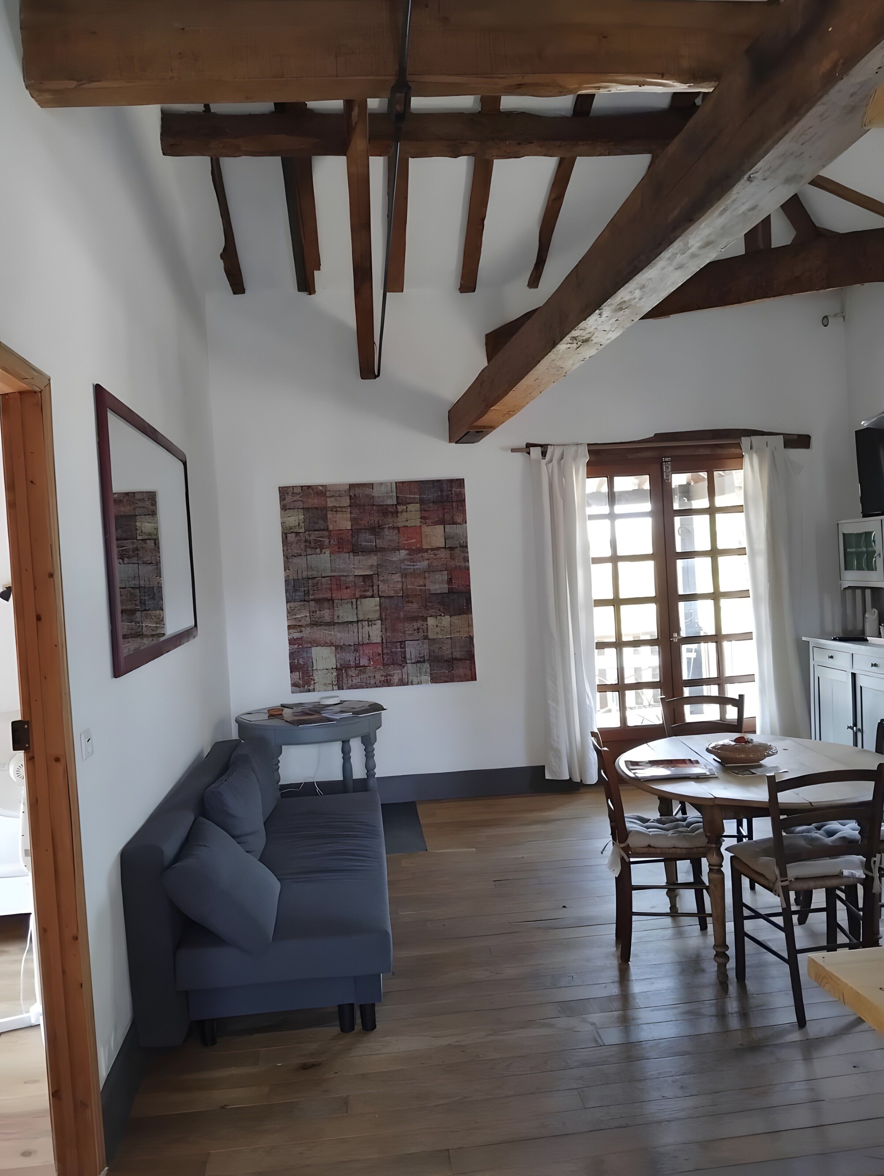 A room with a wooden ceiling, a gray sofa, a dining table with chairs, a window with curtains, and a wall-mounted artwork.