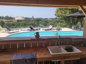 Un espace aménagé au bord de la piscine, avec des chaises longues, une terrasse en bois, un évier et une vue sur un paysage naturel.