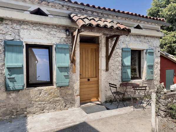 Un bâtiment rustique avec une porte en bois, une fenêtre avec des volets et un petit espace extérieur aménagé pour s’asseoir.