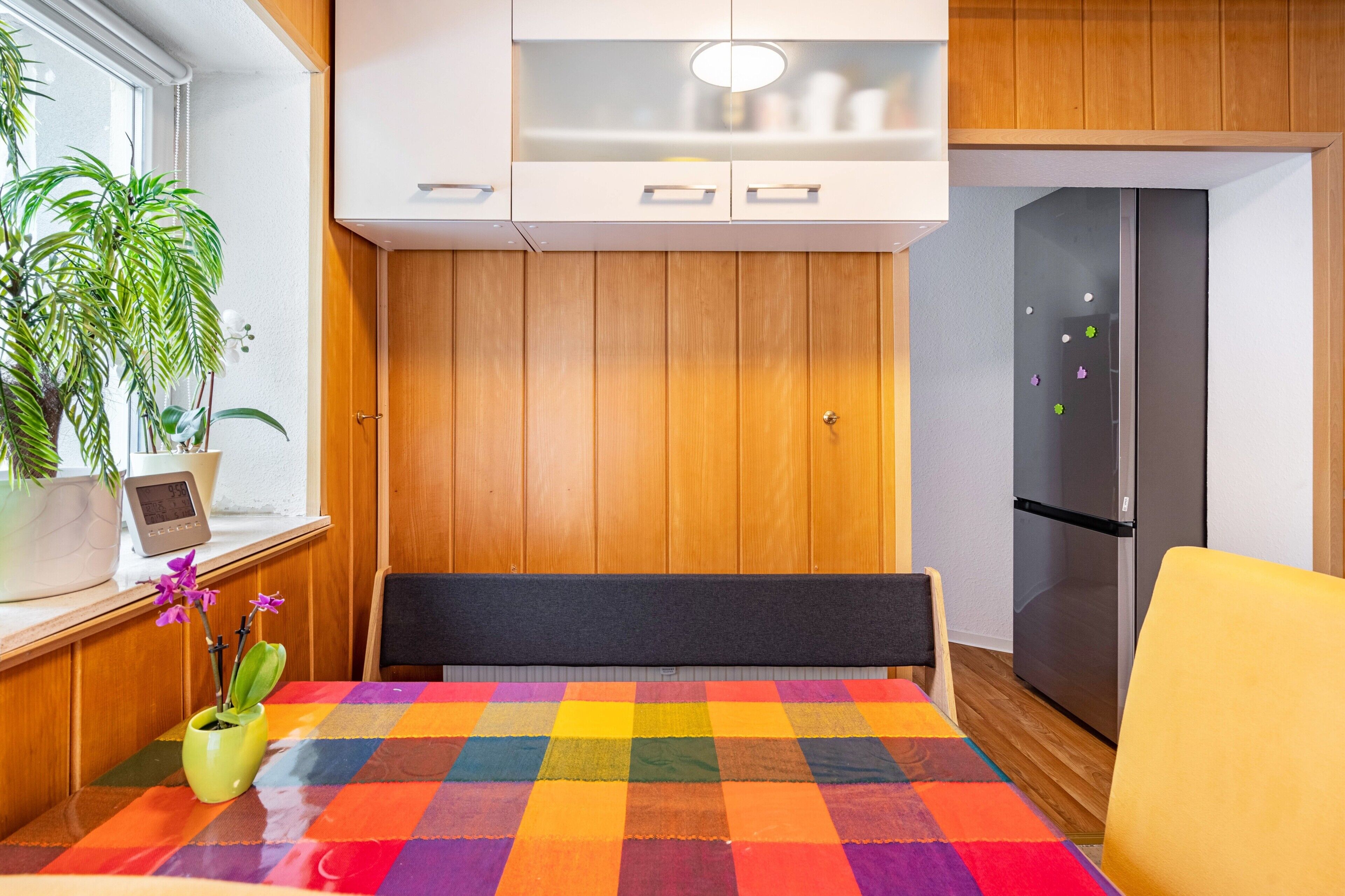 A kitchen with wooden cabinets, a refrigerator, a colorful table, and a potted plant.