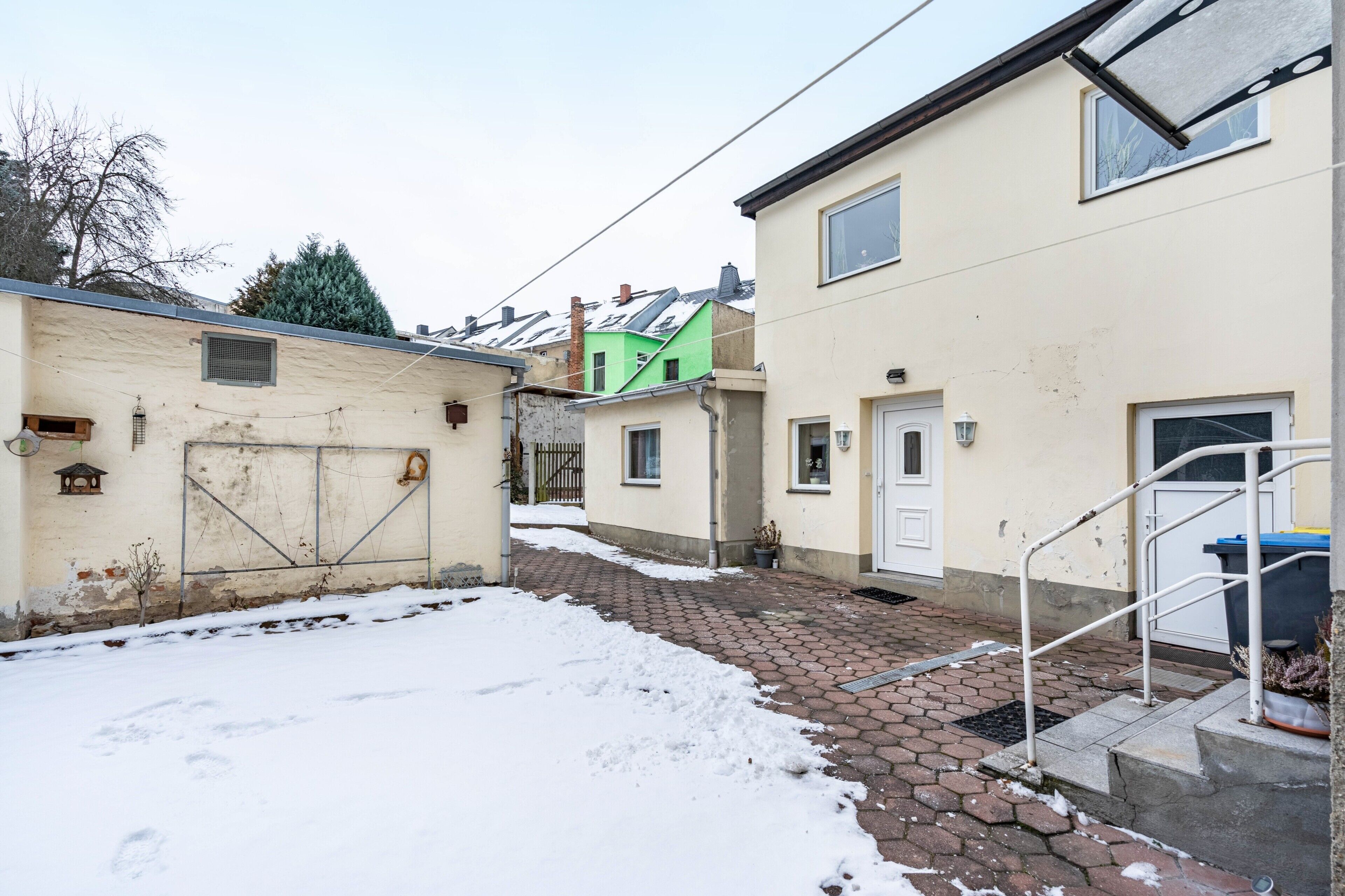 A narrow street with snow-covered ground, a green house, and a beige house with a white door.