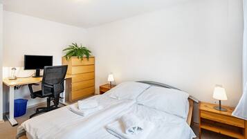 Een hotelkamer met een bed, een bureau met een computer, een stoel, een plant en twee bedlampen.