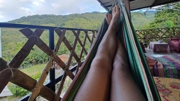 A hammock under a roofed area with a view of green hills.
