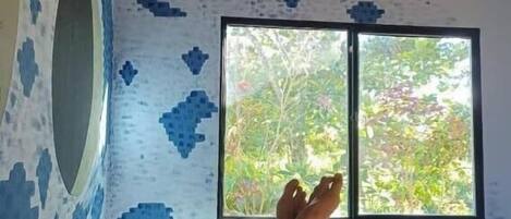 A bedroom with a bed, a window with a view of greenery, and a patterned wall.