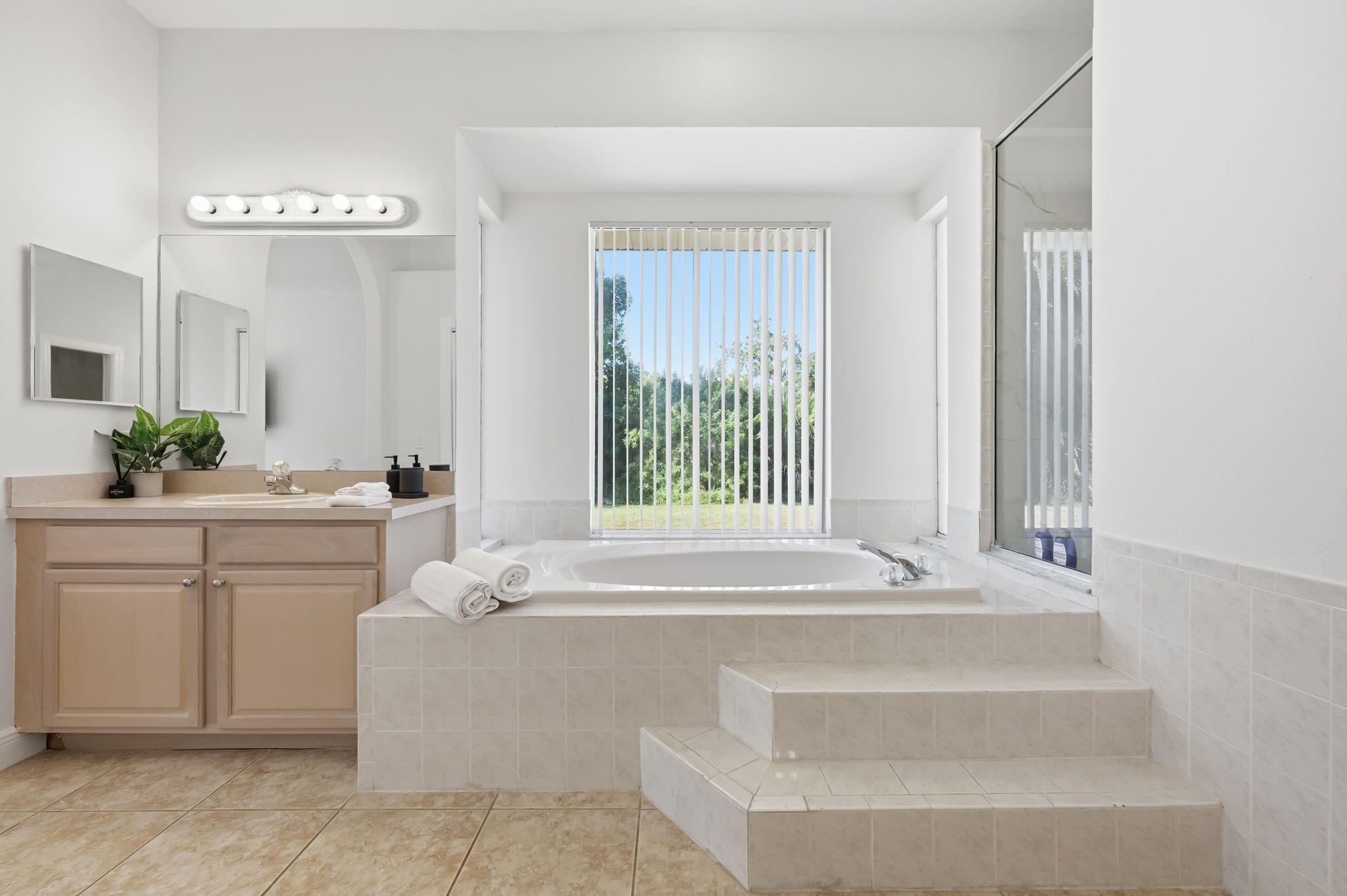 Une salle de bain moderne avec une grande baignoire, un meuble-lavabo avec un miroir et une fenêtre équipée de stores verticaux.