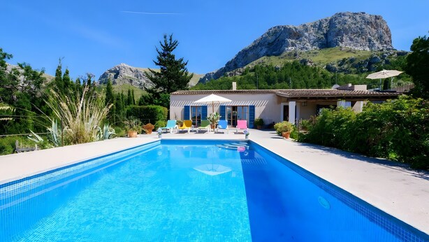 Une piscine avec une vue sur les montagnes, un bâtiment aux volets bleus et des espaces de détente en plein air.