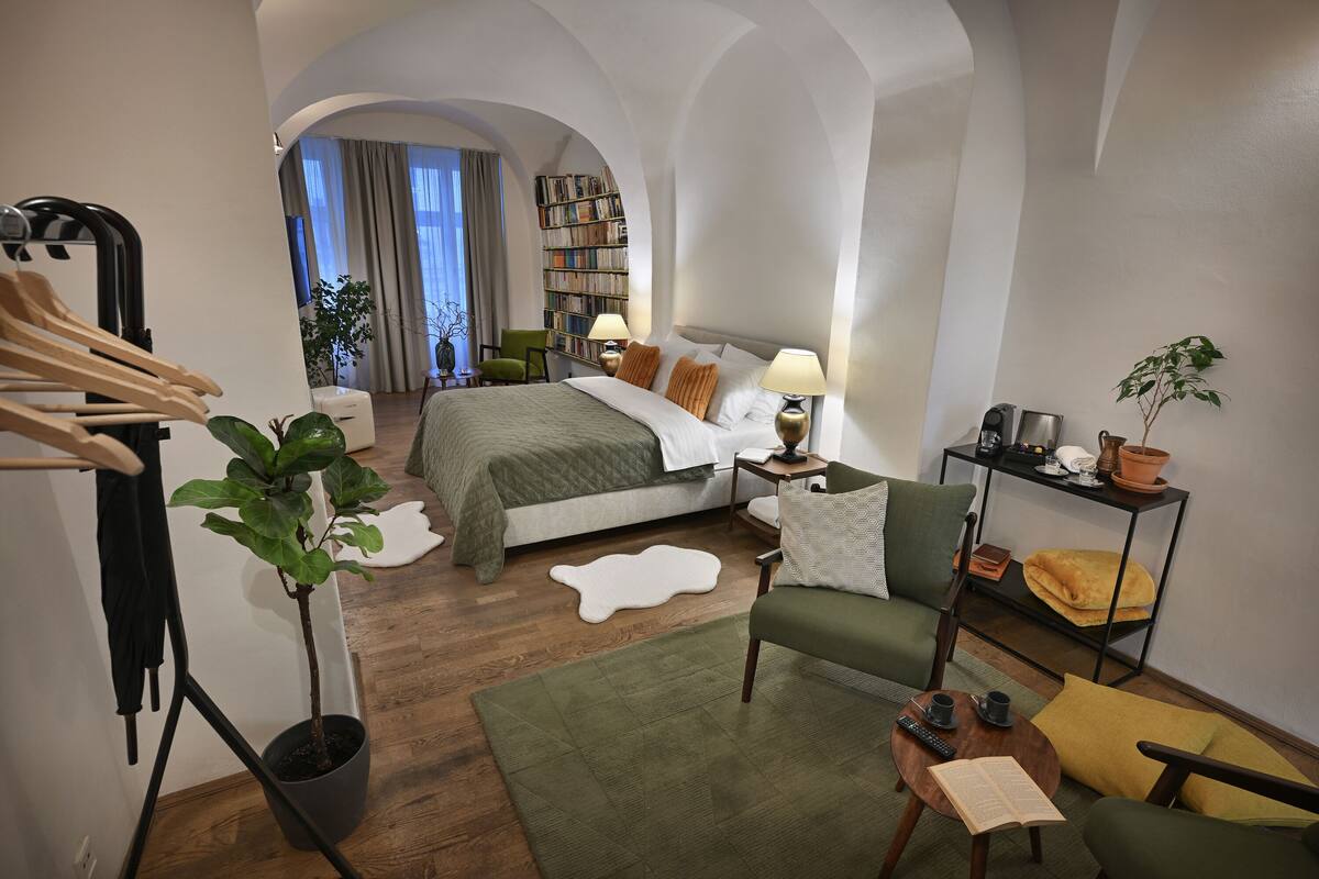 Habitación de hotel con una cama, un sillón verde, una mesita con un libro y una taza, una estantería y una planta en maceta.