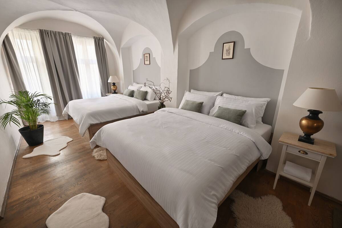 Habitación de hotel con dos camas, una planta en maceta, una lámpara y vistas a través de un ventanal de arco.