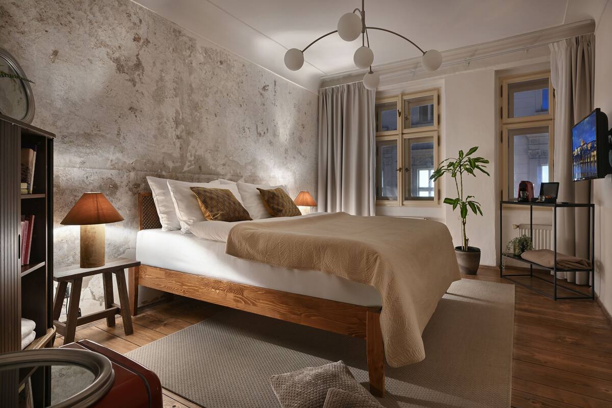 Un dormitorio con una cama grande, cabecera de madera, una mesita de noche con lámpara, una estantería, una planta en maceta y un televisor sobre un mueble.
