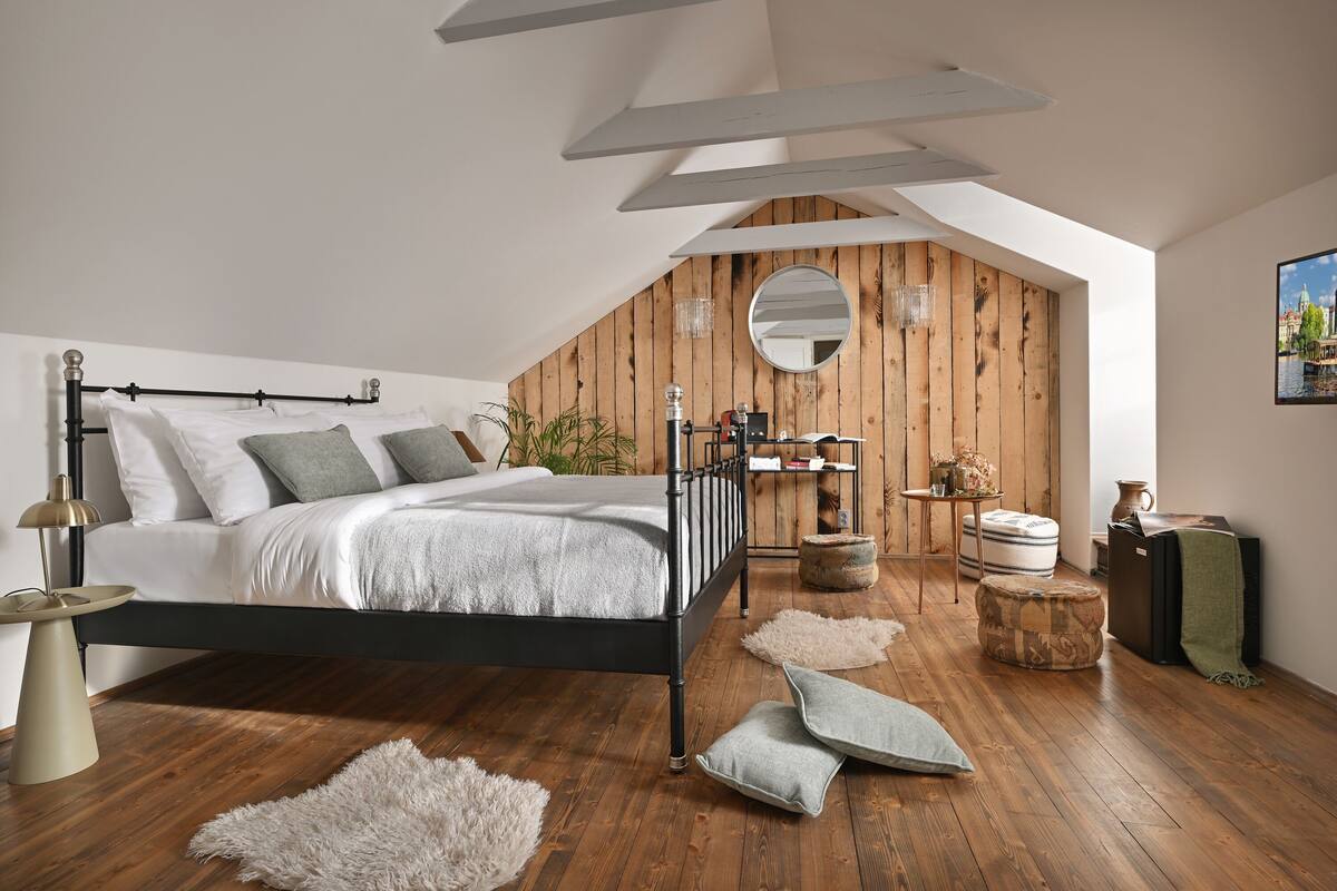 Un dormitorio con techo inclinado, una cama con estructura negra, suelo de madera, un espejo redondo y una mesita con lámpara.