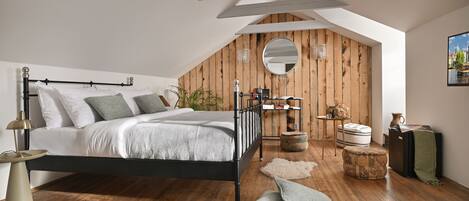 A bedroom with a sloped ceiling, a bed with a black frame, wooden flooring, a round mirror, and a small table with a lamp.