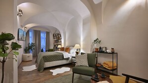 Una habitación de hotel moderna con una cama grande, una zona de estar con un sillón verde, una mesita de centro, una estantería y un ventanal con cortinas.