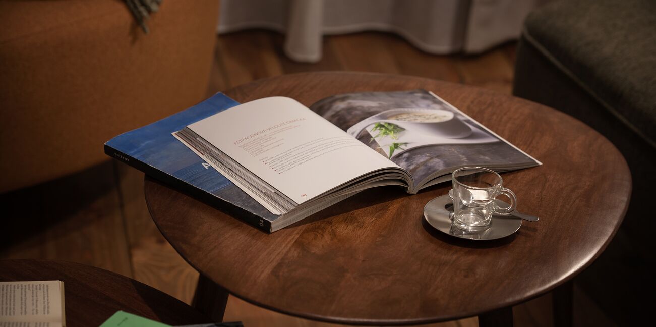 An open book on a small round table with a glass cup on a saucer beside it.