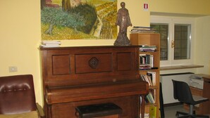 A room with a wooden piano, a bench, a bookshelf, a chair, and a painting on the wall. - Ostello della Pace Assisi (Assisi)