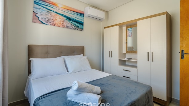 A neatly arranged bedroom with a bed, a wardrobe, and a painting of a beach scene. - Apt w/ 1 Bedroom and Air Conditioning Irn0103 (Florianópolis)