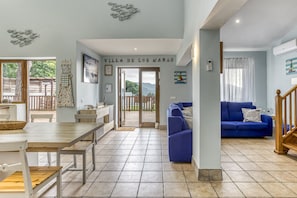 A spacious living area with a blue sofa, a wooden dining table, and a kitchen area visible through an open door. - La Villa de los Mares – Family Villa with Pool and Sea Views on the Costa Brava (Bell-Lloch)