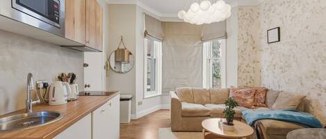 A modern kitchen with a built-in microwave, a sink, and wooden cabinets. There is a dining area with a round table and chairs, a sofa, and a decorative plant.